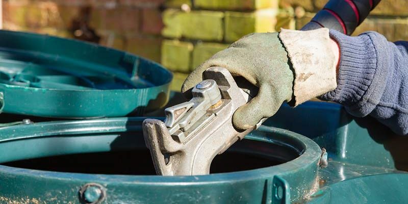 Gloved hand refuelling an oil tank with a fuel nozzle outdoors near a brick wall.